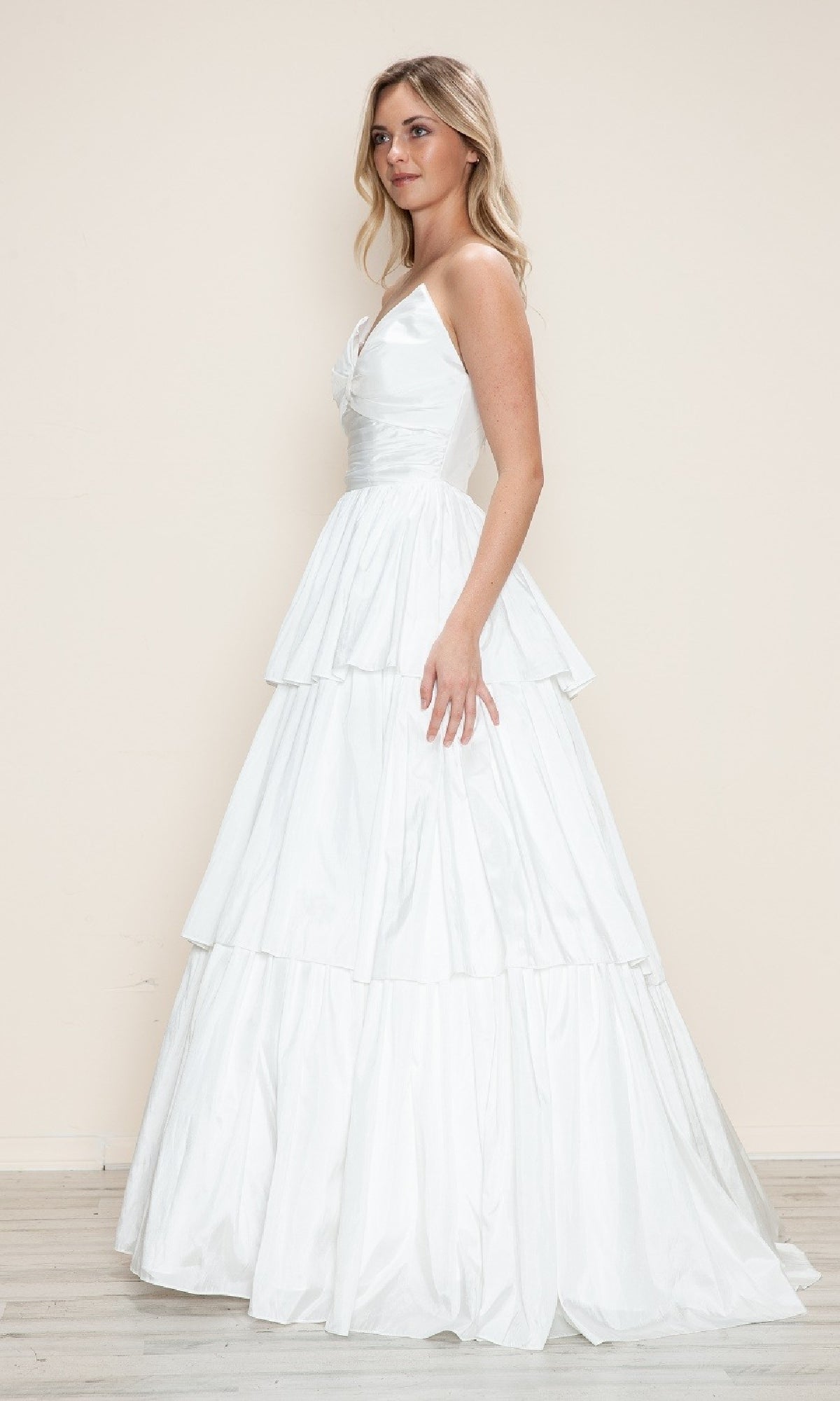 A woman stands indoors wearing the Long Prom Dress: Poly USA 9694, a white, floor-length gown with a fitted bodice and tiered a-line skirt.